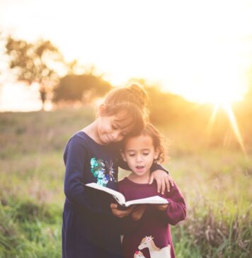Confie os filhos a Cristo girl's left hand wrap around toddler while reading book during golden hour