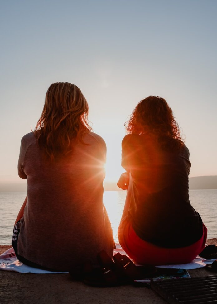 Photo by Briana Tozour two woman sitting on beach sand while facing sunlight