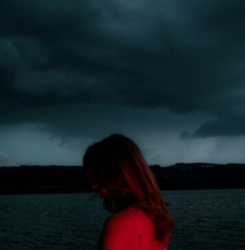 Onde a graça abunda A woman in a light dress illuminated by red light against a dark lake