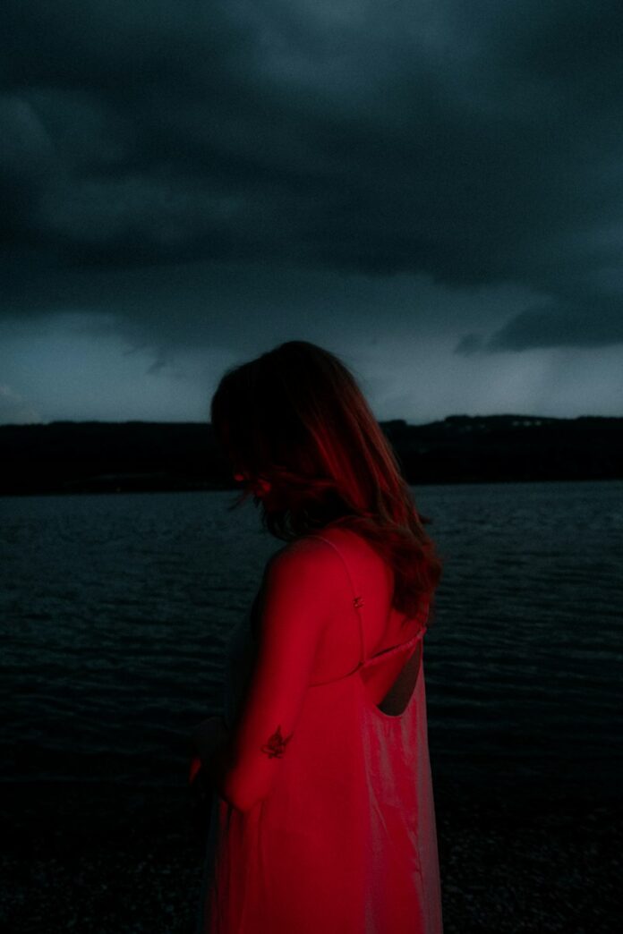Photo by Elijah Grimm A woman in a light dress illuminated by red light against a dark lake